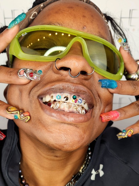 Playful portrait in green sunglasses showing a full row of colorful crystal tooth gems with gold accents