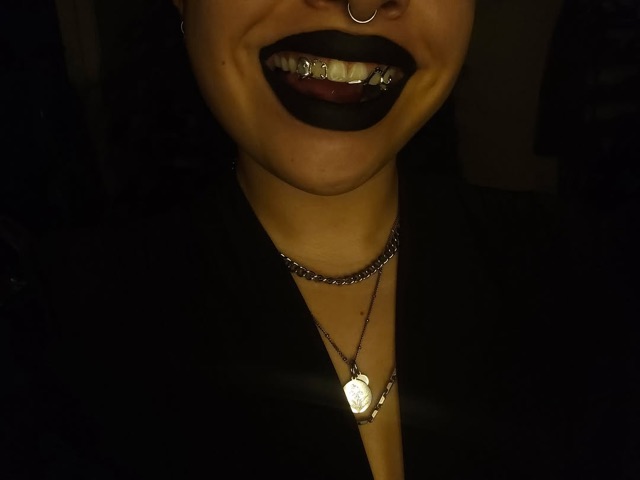 Moody low-light portrait with black lipstick showing open-design grillz on the upper teeth