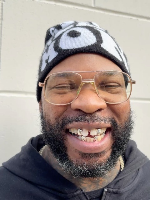 Smiling client in a beanie and aviator glasses showing a custom gold-and-stone grillz set on the upper front teeth