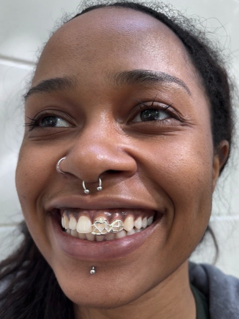 Smiling client with nose and labret piercings showing a linked white-gold infinity design across the upper front teeth