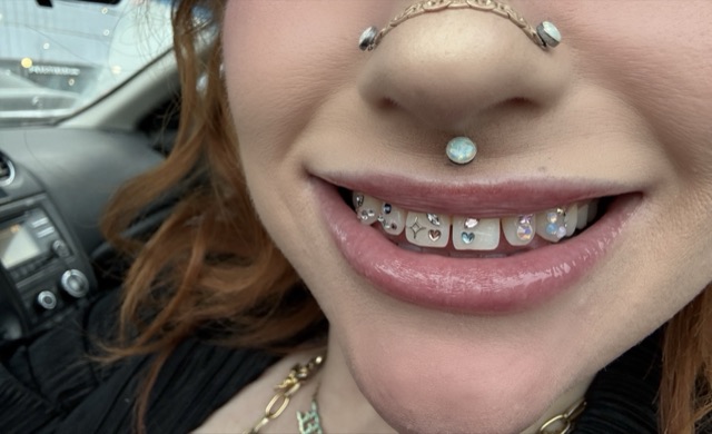 Close-up of a full row of crystal tooth gems, including a star and two hearts, worn with an opal septum ring and nose stud