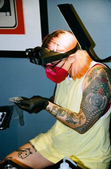 Gemmy in a red respirator mask, magnifying loupe headset and black gloves, preparing jewelry at the studio bench