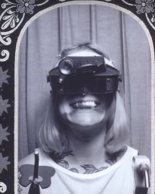Black-and-white photobooth portrait of Gemmy grinning in a magnifying loupe headset, framed by the booth's ornate arch